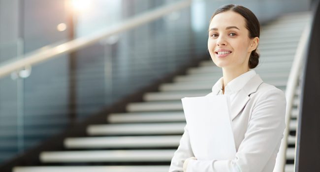 Young agent with papers standing by staircase in airport lounge and looking at camera