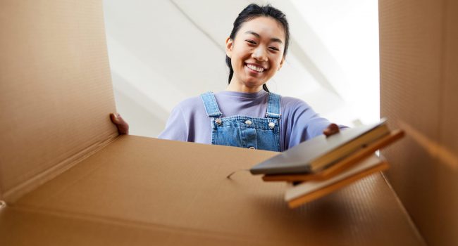 Low angle view at young Asian woman looking into cardboard box and smiling happily while packing or unpacking for new home, copy space