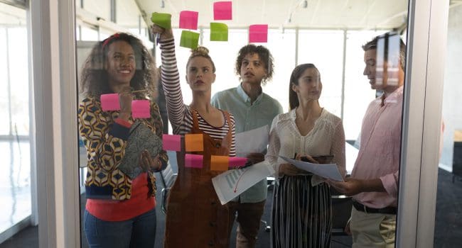 Front view of multi-ethnic business colleagues discussing business strategy over sticky notes in modern office Front view of multi-ethnic business colleagues discussing business strategy over sticky notes in modern office