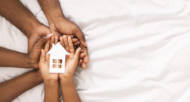 Adoption and family housing concept. Cutout paper house in hands of african american family over white bed sheet background. Closeup, panorama