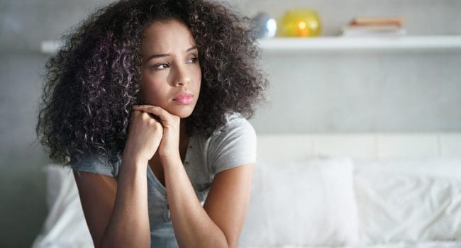 Lonely young latina woman sitting on bed. Depressed hispanic girl at home, looking away with sad expression.