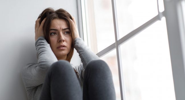 Despaired sad european millennial woman suffering from depression and psychological problems, holding her head near window. Reaction to problems, depression and tension during covid-19 quarantine