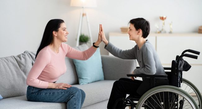 Happy teenage boy in wheelchair high fiving his mother at home, side view. Cheerful disabled adolescent and his parent celebrating achievement or success together, indoors