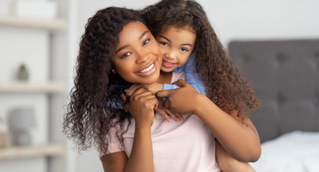 Love between parent and child. Positive little black girl hugging her mom on bed at home. Young African American woman and her daughter embracing, spending quality family times together indoors