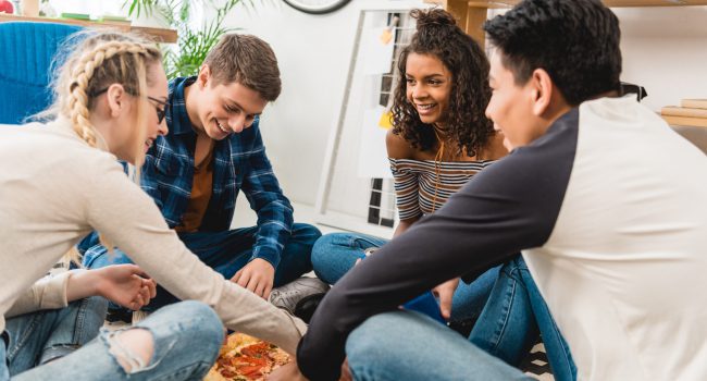 multiethnic teen friends sitting on floor and eating pizza