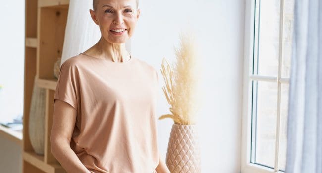 Positive beautiful mature woman in casual wear keeping hands in jeans pockets and smiling at camera Portrait of attractive happy middle aged woman in casual wear keeping hands in jeans pockets and smiling at camera while standing near window indoors. Positive senior people and lifestyle concept