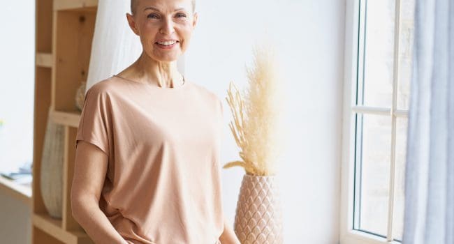 Portrait of attractive happy middle aged woman in casual wear keeping hands in jeans pockets and smiling at camera while standing near window indoors. Positive senior people and lifestyle concept