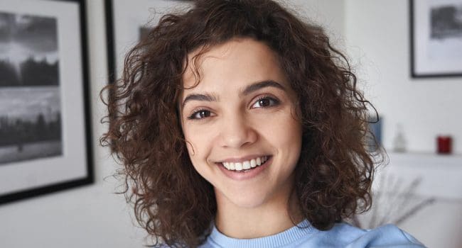 Smiling cute cheerful young pretty hispanic latin woman looking at camera standing alone at home. Happy positive beautiful 20s girl female model posing indoors, close up face headshot portrait.