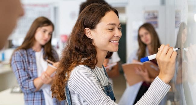 Happy college student writing equation on white board in class. Satisfied young girl solving math problem on whiteboard with classmates in background watching her. Proud high school student smiling while writing during a math class.